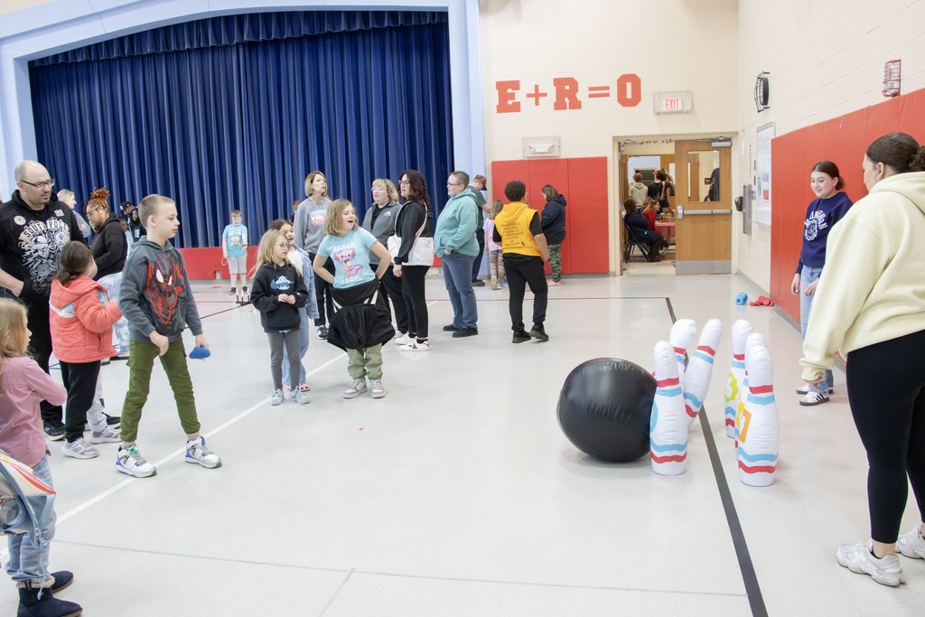 A boy rolls a giant bowling ball and hits giant blow up bowling pins while people watch