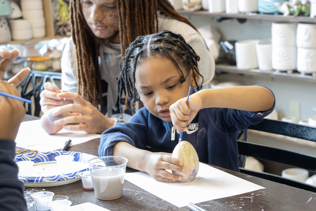 A young child sits at a table painting a ceramic egg with a small brush, concentrating carefully on the details. An adult with long hair sits just behind them, assisting and guiding the activity. The table is covered with paint supplies, including cups of water, paint trays, and paper plates. Shelves filled with pottery pieces are visible in the background, suggesting a ceramics or art studio setting.