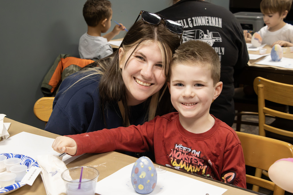 A smiling adult and young child sit together at a table in an art studio, posing for the camera. The child holds a painted ceramic egg decorated with colorful dots. Paint supplies, including small cups and brushes, are spread across the table. In the background, other children are also painting, creating a lively, creative environment.