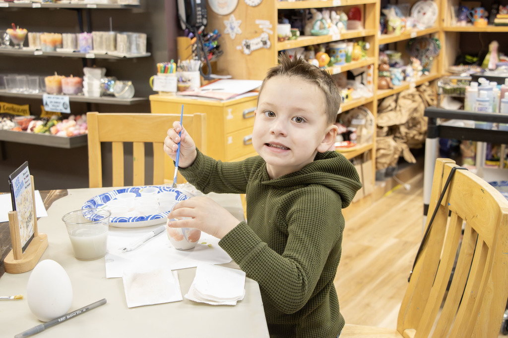 A young child sits at a table in a pottery studio, holding a paintbrush and working on a ceramic egg. The child looks toward the camera with a slight smile while painting. The table has paper towels, a cup of water, and paint supplies. Behind them, shelves are filled with various pottery pieces and art materials, creating a colorful, creative studio setting.