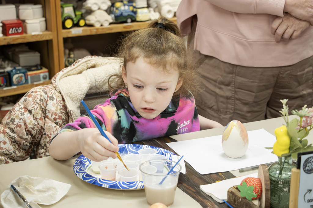 A young child sits at a table in a pottery studio, carefully dipping a paintbrush into small cups of paint arranged on a paper plate. A partially painted ceramic egg sits nearby on a sheet of paper. The child appears focused on choosing colors, while an adult stands nearby. Shelves filled with ceramic items and art supplies are visible in the background.