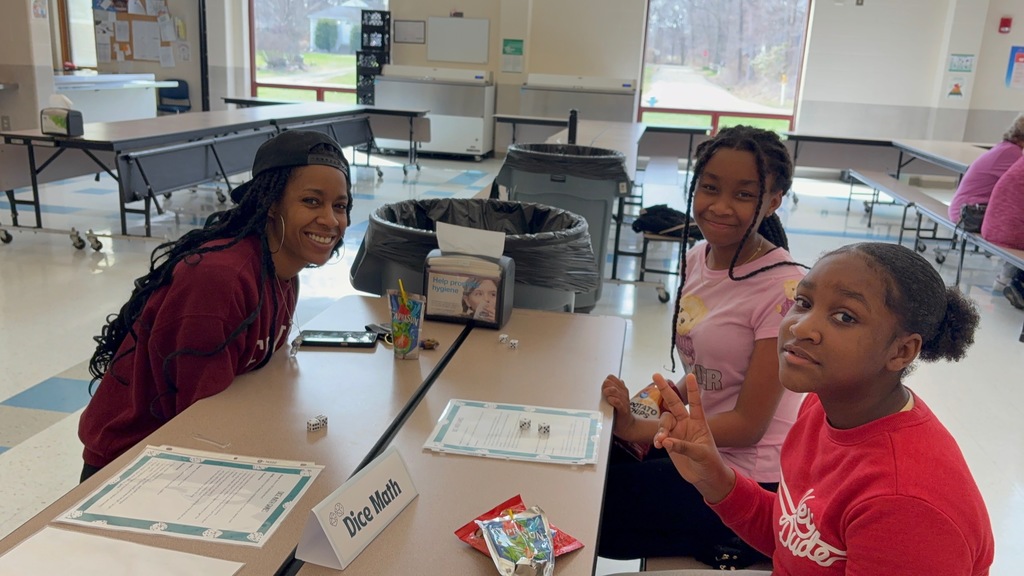 An adult and two students sit at a cafeteria table playing a math activity labeled “Dice Math.” All three smile at the camera, with one student holding up a peace sign. Dice, worksheets, and snacks are spread across the table, and the cafeteria space is visible in the background.