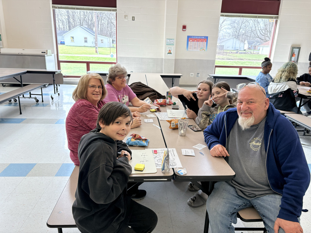 A group of students and adults sit together at a cafeteria table working on a math activity. Several smile at the camera, and two students make peace signs. Papers, small game pieces, and snacks are spread across the table, with large windows and additional groups visible in the background.