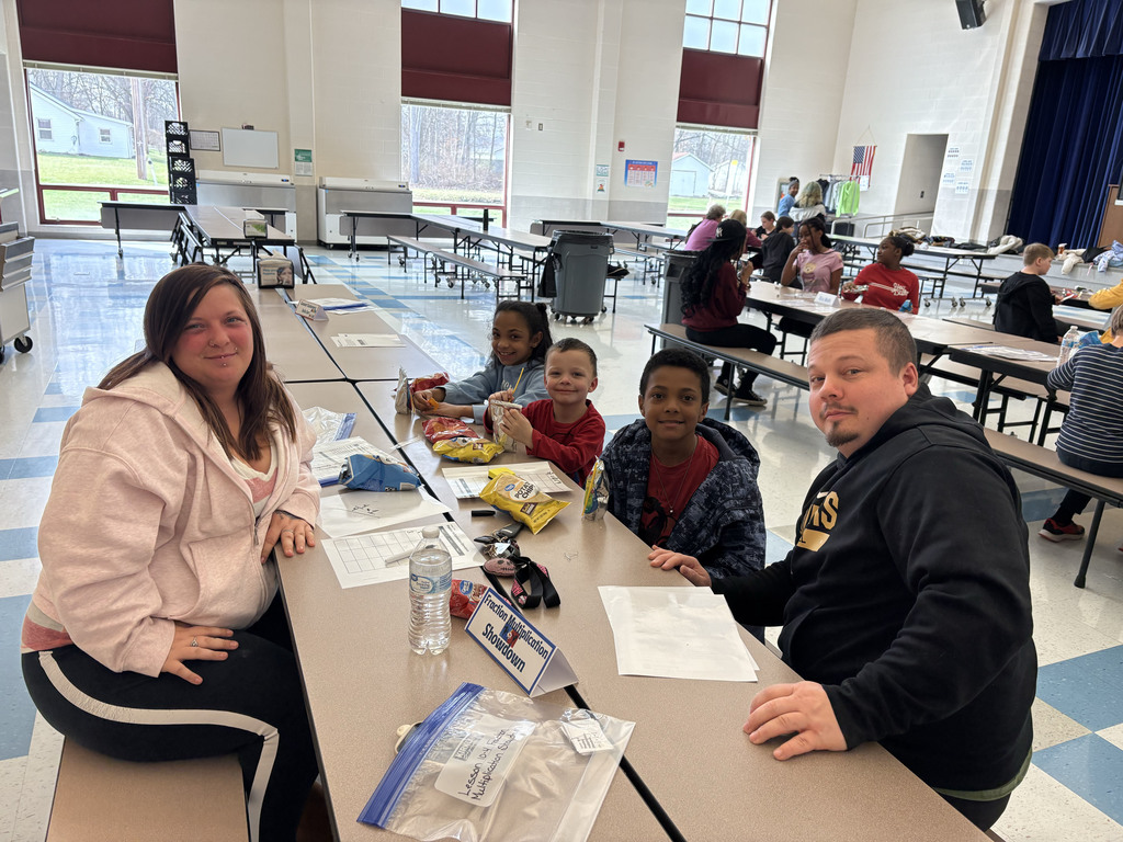 Two adults and three students sit together at a cafeteria table participating in a math activity labeled “Fraction Multiplication Showdown.” They smile at the camera with worksheets, snacks, and water bottles on the table. Other families and students are gathered at tables in the background of the bright cafeteria.