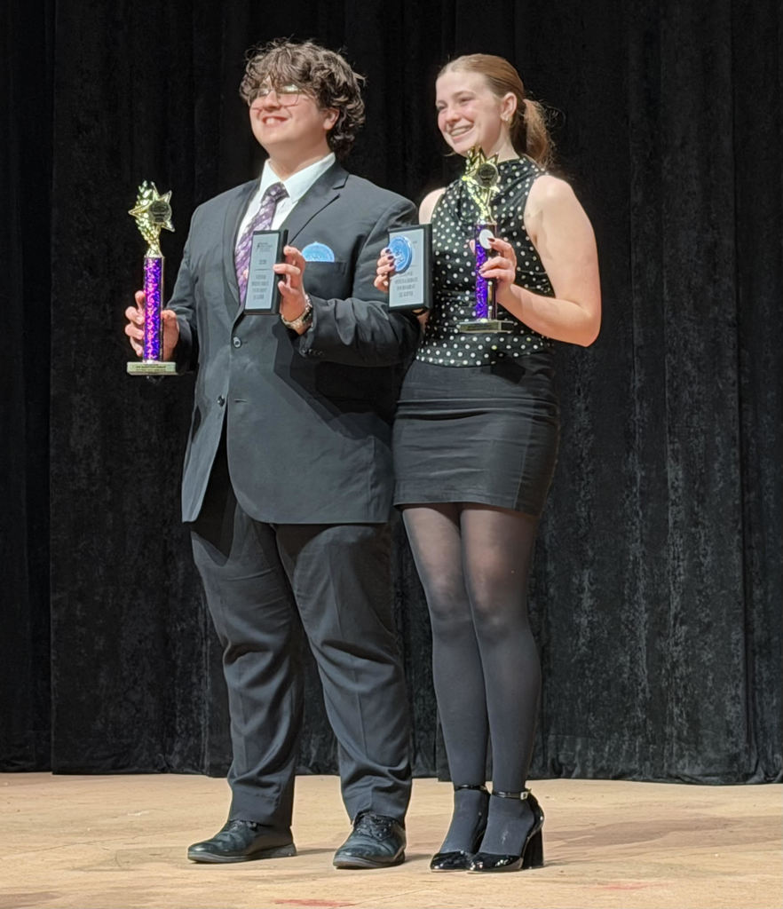 Aaden Vega standing on stage with another winner holding a trophy and plaque while smiling