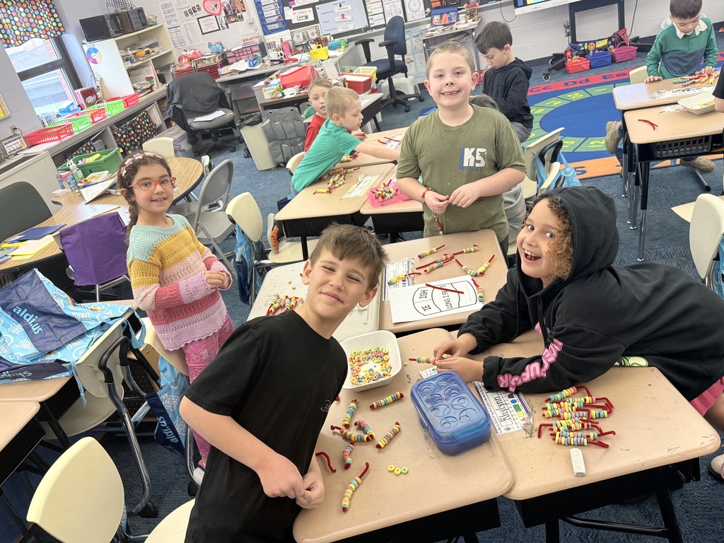 A group of elementary students smile and work together at their desks, using colorful cereal and pipe cleaners as hands-on math tools. The classroom is bright and busy, with students collaborating, building, and learning through interactive activities.