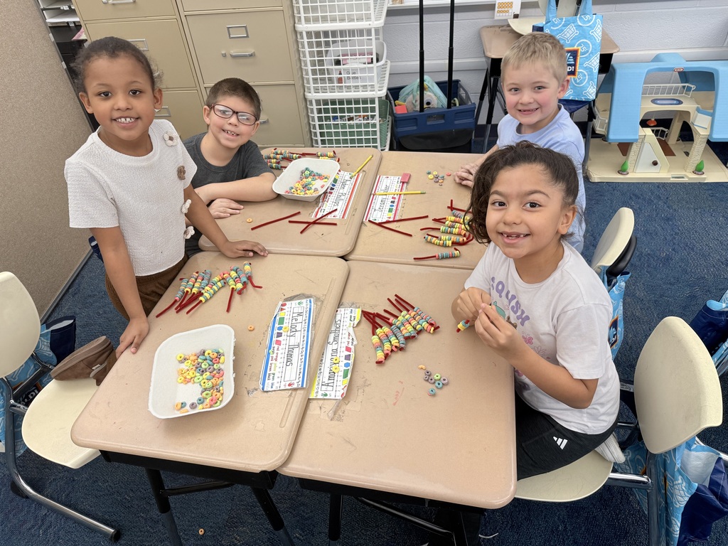 Four elementary students sit together at a cluster of desks, smiling at the camera while using colorful cereal and pipe cleaners to build hands-on math models. Bowls of cereal and classroom materials are spread across the tables as they work and collaborate in a bright classroom setting.
