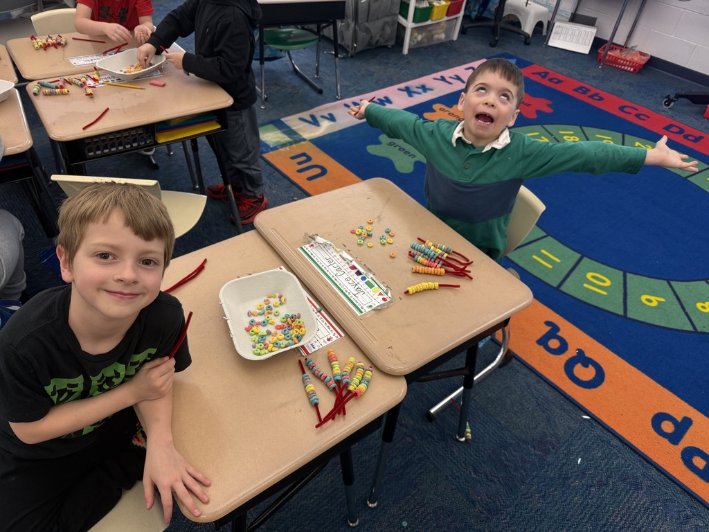 Two elementary students sit at their desks working with colorful cereal and pipe cleaners for a hands-on math activity. One student smiles at the camera while the other playfully throws his arms up, showing excitement. A bright classroom rug and additional students working in the background complete the lively learning environment.
