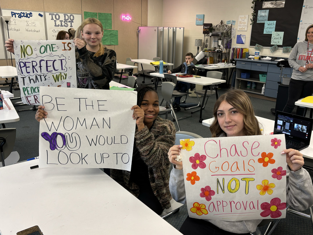 Students holing up positive posters they made that say "Be the woman you would look up to," No one is perfect that's why pencils have erasers," and "Chase goals not approval"
