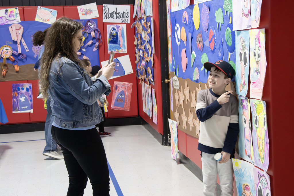 A student stands next to a colorful wall of artwork, smiling and pointing to their piece while a family member takes their photo on a phone. The display features a variety of bright, creative student projects, and a sign above reads “Mrs. Robertson’s class.”