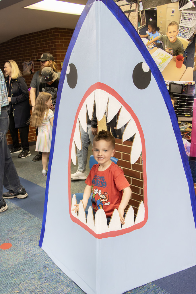 A young child smiles while sitting inside a large, painted shark display with an open mouth, designed for photos. The shark structure is light blue with white teeth and a red outline around the mouth. In the background, families and children stand and talk in a school hallway, and a small collage of classroom photos is displayed on the wall.