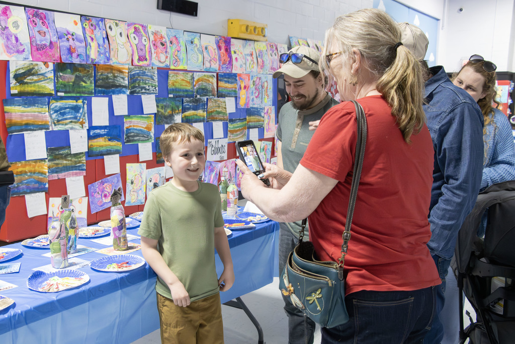 A student stands smiling in front of a colorful display of artwork while a family member takes their photo on a phone. The wall behind them is filled with bright paintings and drawings, and a table below showcases additional student projects. Other family members stand nearby, looking on during the school art event.