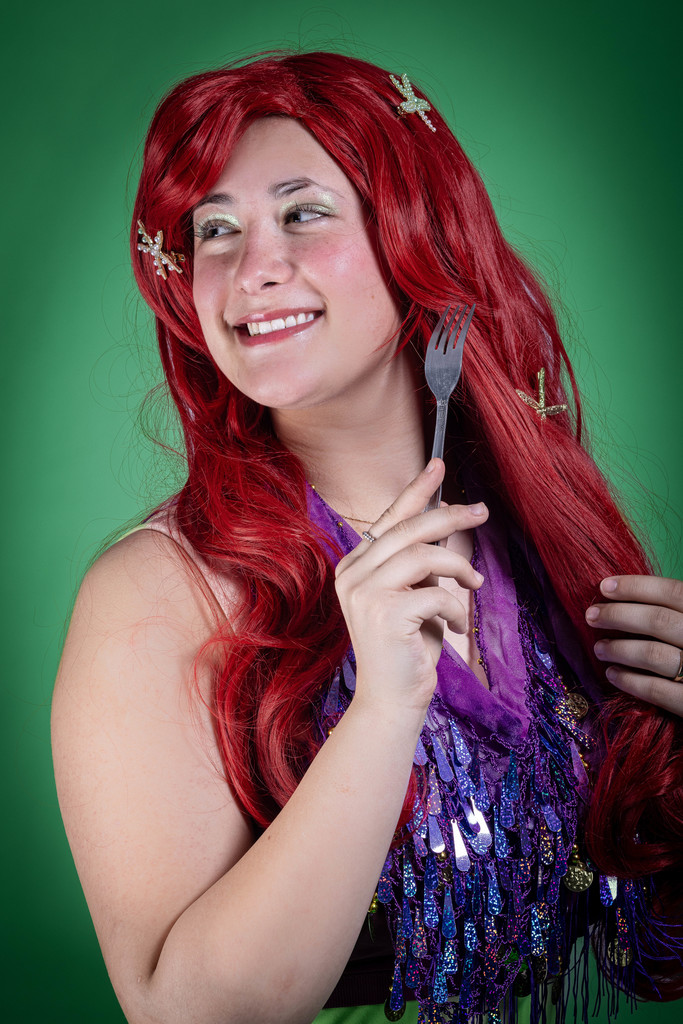 A student dressed as Ariel from The Little Mermaid smiles while holding a fork, referencing the character’s “dinglehopper.” They wear a long red wig with starfish clips and a purple costume top against a green backdrop.