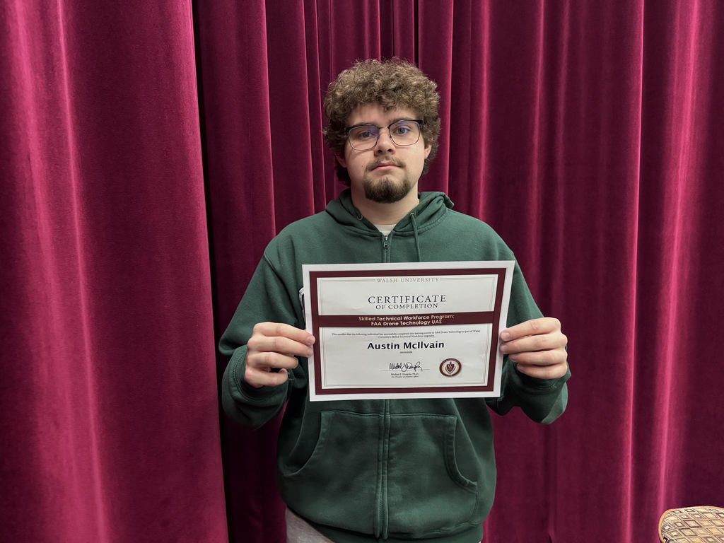 A student stands in front of a red curtain holding a certificate of completion from Walsh University. The certificate recognizes Austin McIlvain for completing the Skilled Technical Workforce Program: FAA Drone Technology UAS.