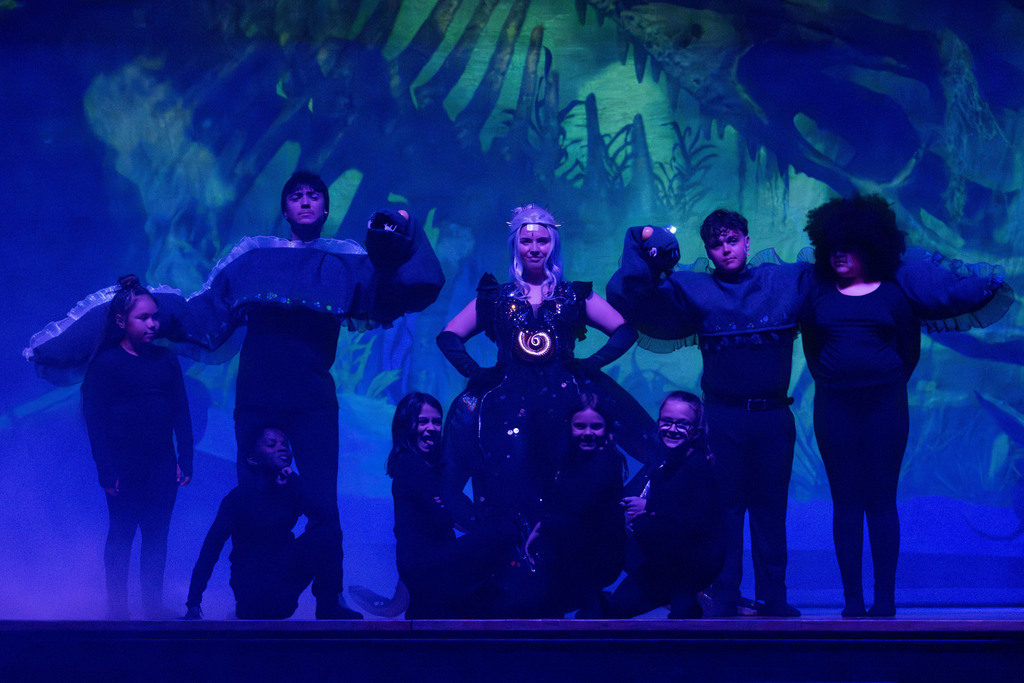 A group of students performs on stage under blue lighting in an underwater-themed scene. At the center, a student dressed as Ursula from The Little Mermaid stands confidently in a black costume with a glowing shell necklace. Surrounding them are other students in black outfits, some wearing large sea creature costumes resembling eels, posed with arms raised. Additional students kneel in front, smiling. The backdrop features ocean imagery, creating a dramatic theatrical setting.