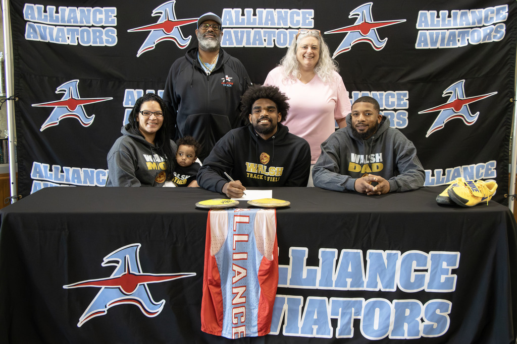 An Alliance High School student-athlete signs to continue track and field at Walsh University, surrounded by family. The student sits at a table in front of an “Alliance Aviators” backdrop, wearing a “Walsh Track & Field” sweatshirt and smiling while signing. Family members stand and sit around him, including individuals wearing “Walsh Mom” and “Walsh Dad” sweatshirts, along with a young child. On the table are track and field items, including two discus and a pair of yellow track shoes. An Alliance uniform hangs over the front of the table, highlighting the athlete being celebrated.