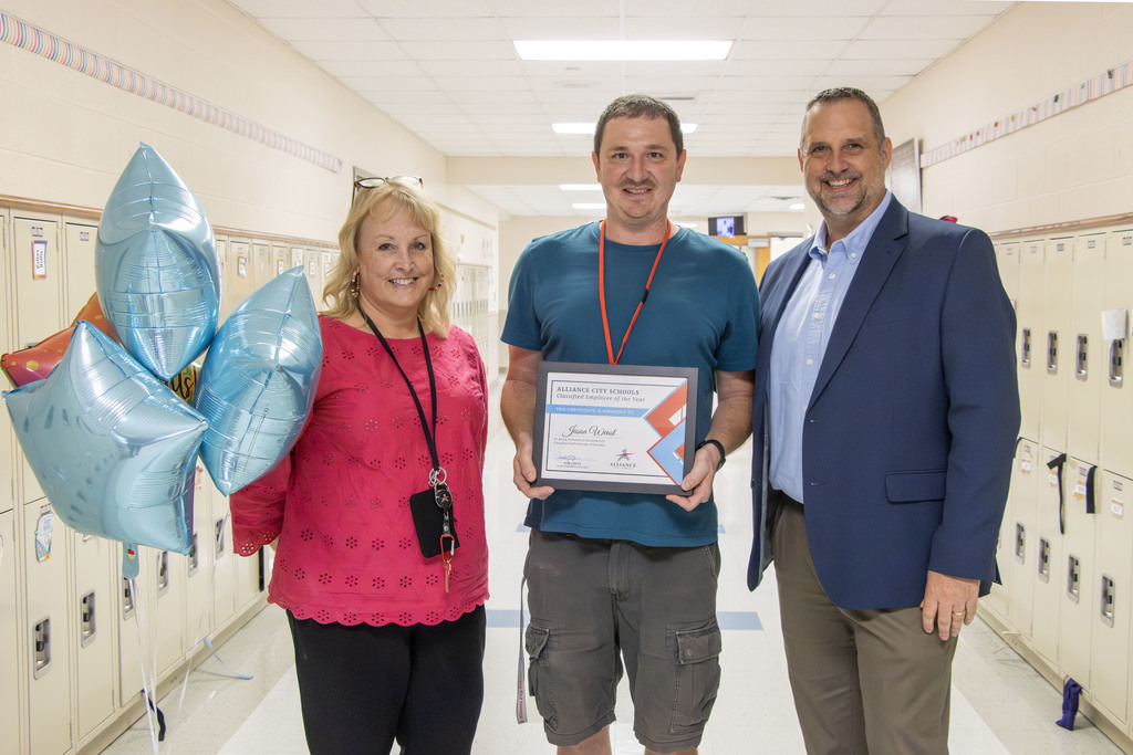 Three people stand in a school hallway with lockers in the background. A woman on the left holds a bouquet of blue and red balloons.