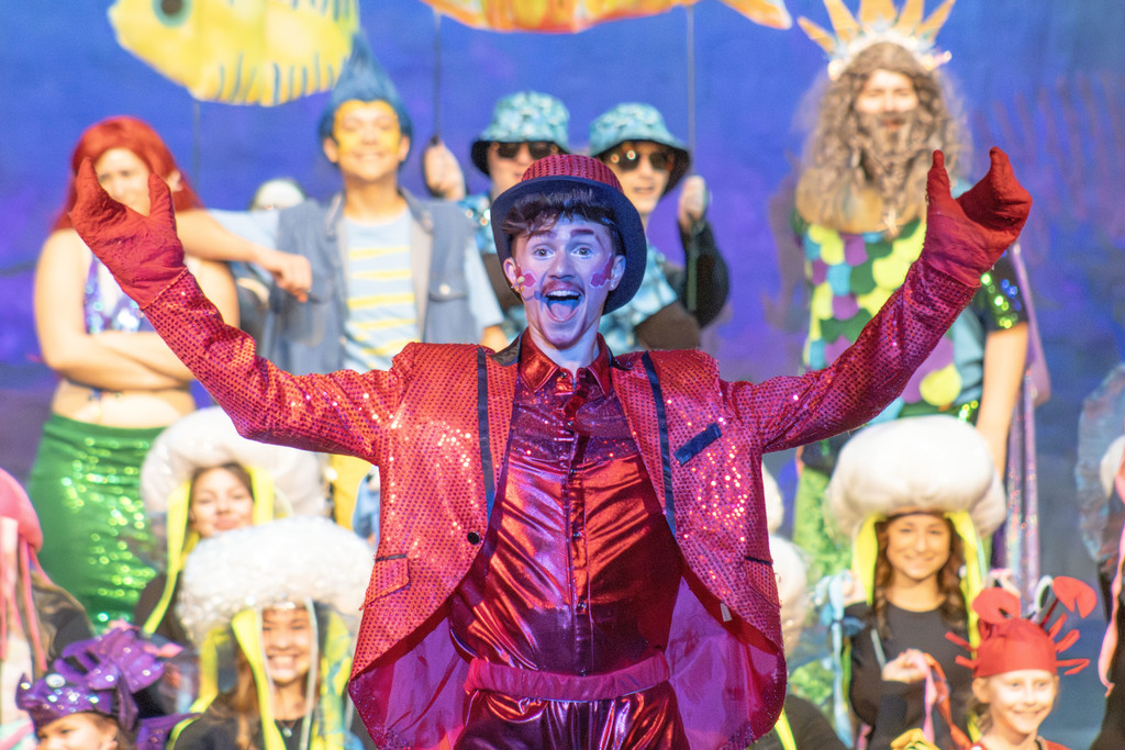 A high school theater performance scene featuring a student in a bright red, sequined costume and top hat standing center stage with arms raised and a big smile. Behind them, other students in colorful sea-themed costumes, including fish, sea creatures, and mermaid-inspired outfits, pose under vibrant stage lighting, creating an energetic underwater-themed production.