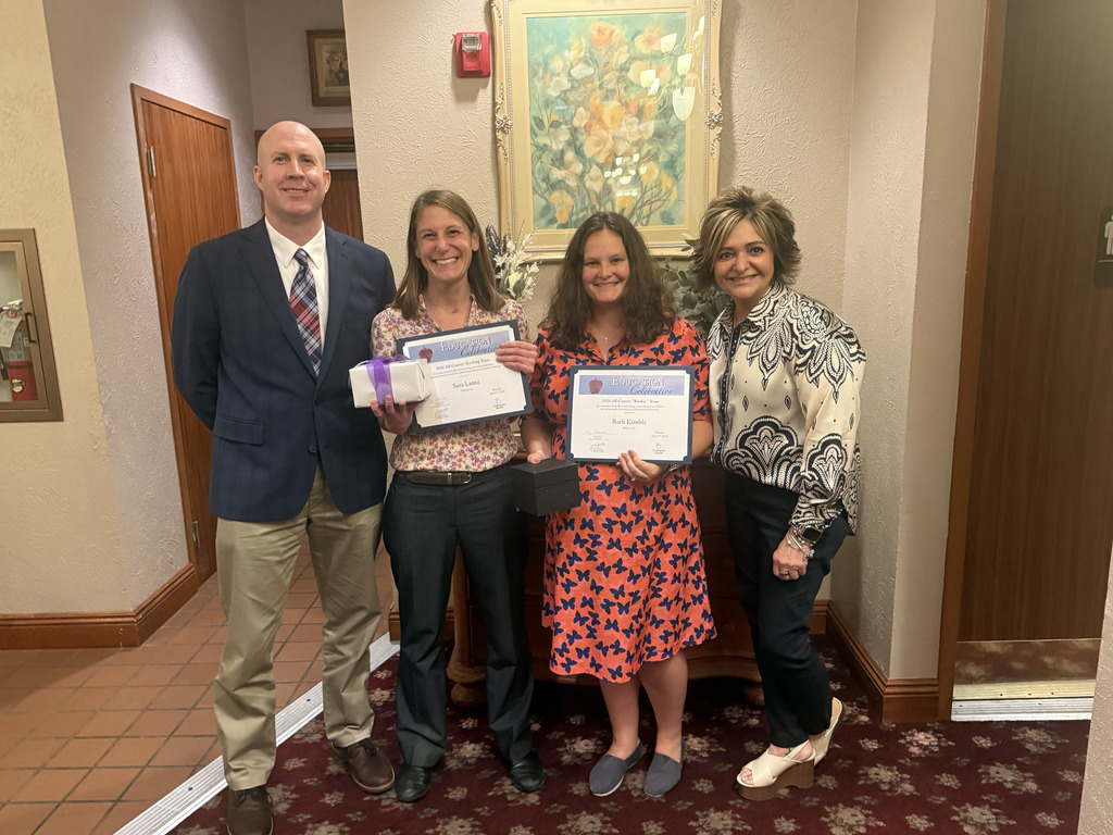 Four adults stand together indoors, smiling and posing for a photo. Two women in the center hold certificates and small gift boxes, appearing to be recognized for an achievement or award. The group is dressed in business attire, standing in a hallway with framed artwork on the wall behind them.