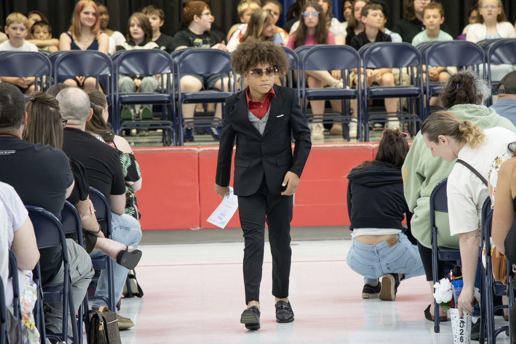A student in a sharp three piece suit walks down the aisle with his certificate in hand