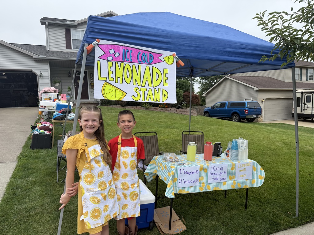 Cora and Henry wearing aprons with lemons on them and standing in front of their lemonade stand.