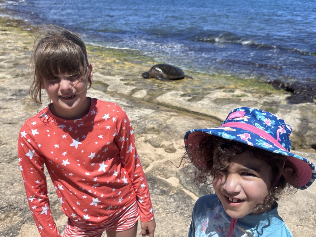 Ella and Emi smiling while sea turtles lay on the beach in the distance behind them