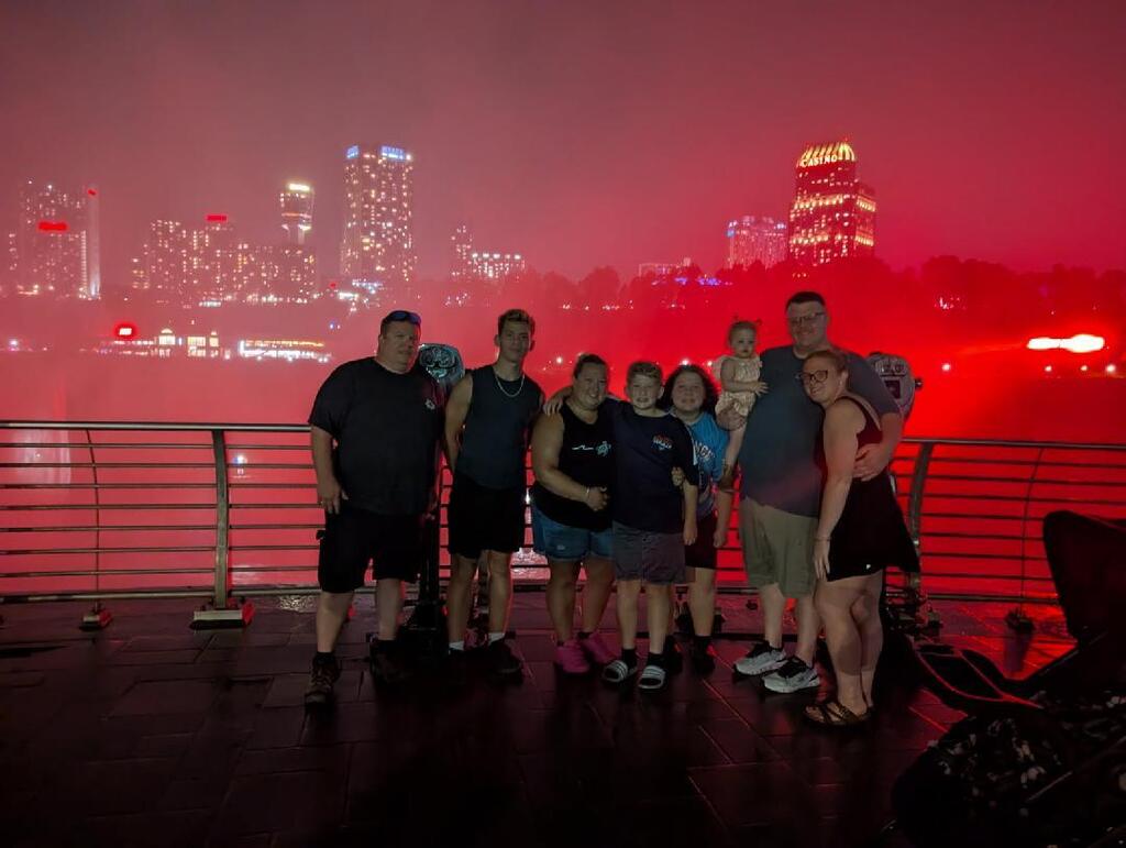 The schuette family standing in front of the falls with the city in the background. The falls are lit up red.
