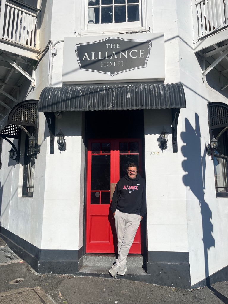 Jason Powers standing at the entrance of a white building with a red door and a sign that says "The Alliance Hotel"