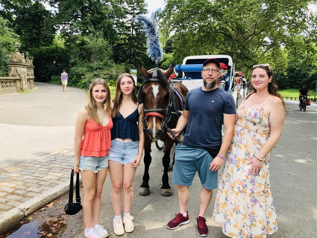 The girls and two older adults posing for a picture with a horse pulling a carriage in a park