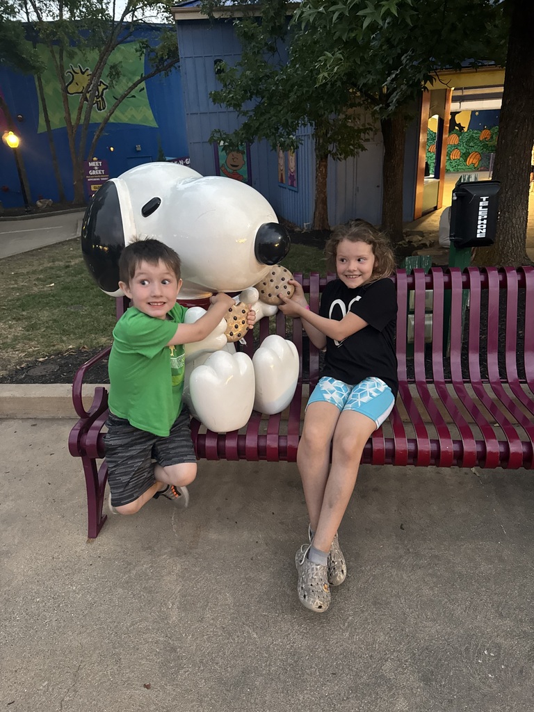Two kids sitting on a bench with a statue of snoopy