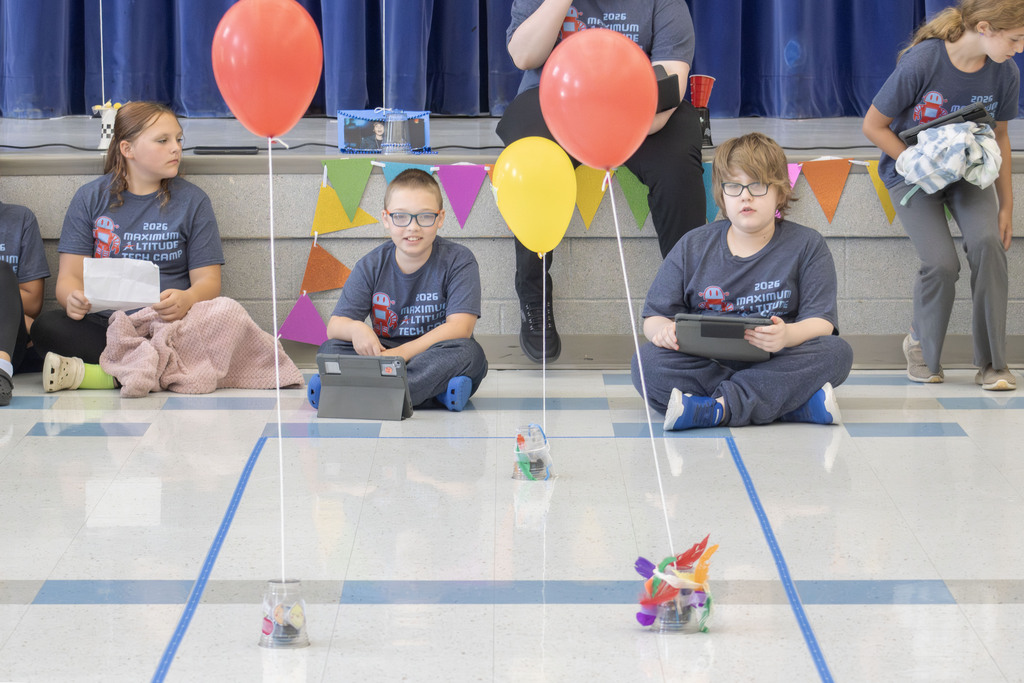Students participating in the Maximum Altitude Tech Camp sit on the floor using tablets to code small robotic parade floats. Three decorated robots are positioned in taped lanes, each topped with colorful balloons and handmade decorations as students test their creations. Other students and staff are visible in the background on the stage, which is decorated with colorful pennant banners. The activity is part of the camp's end-of-week family celebration, where students demonstrated coding and engineering skills by programming their robots to follow a parade route.