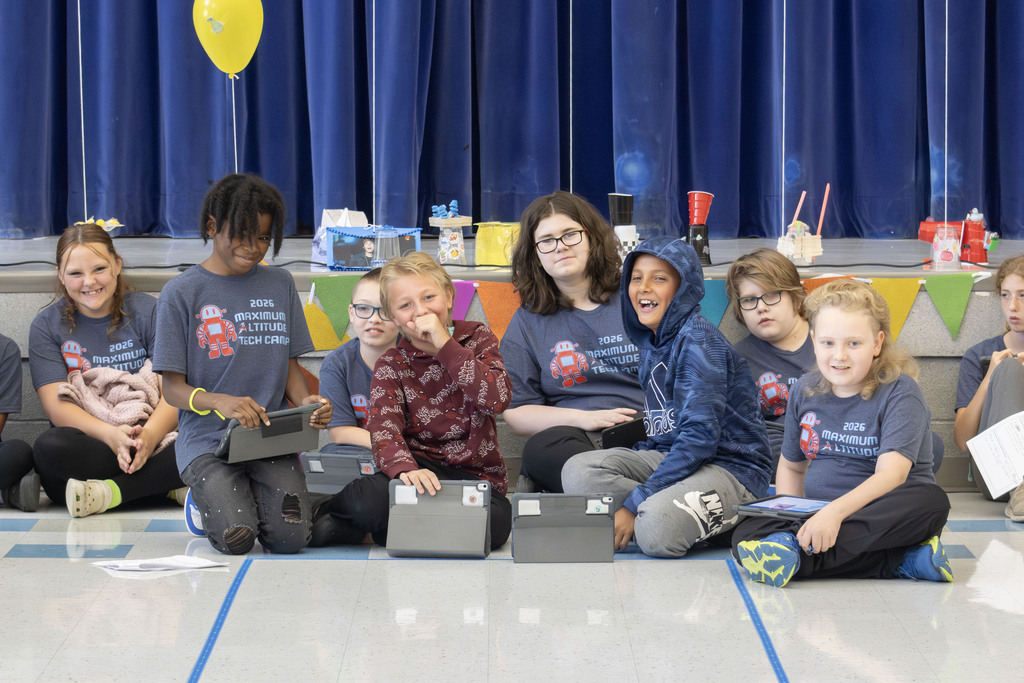 Students participating in the 2026 Maximum Altitude Tech Camp sit together at the starting area of a taped course while holding tablets used to code Sphero Mini Robots. Several students smile and laugh as they wait for their turn to demonstrate their robotic parade floats. The stage behind them is decorated with colorful pennant banners, balloons, and student-built robot floats, highlighting the end-of-week family celebration where students showcased the coding and engineering skills they developed during the camp.