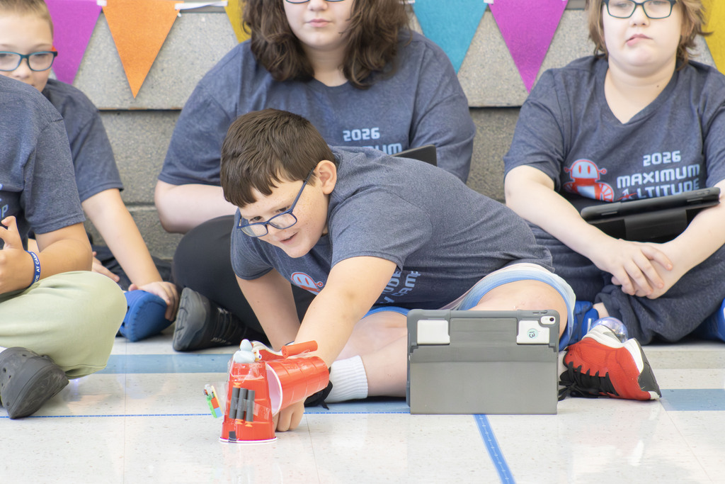 A student participating in the Maximum Altitude Tech Camp kneels on the floor while placing a coded robotic parade float at the starting line of a taped course. The robot is decorated with colorful cups and craft materials, and a tablet used for coding sits nearby. Other students wearing 2026 Maximum Altitude Tech Camp T-shirts watch from behind while holding tablets. Colorful pennant banners decorate the stage in the background as students prepare to demonstrate the coding and engineering skills they learned during the week.