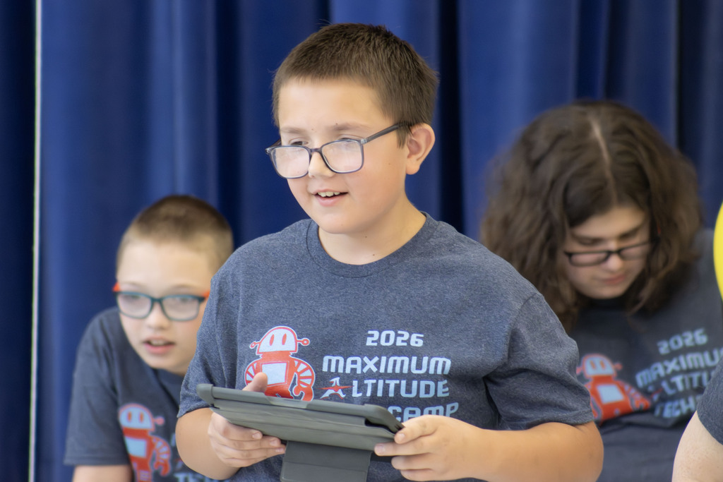 A student smiling while holding an ipad with a shirt that says "2026 Maximum Altitude Tech Camp"