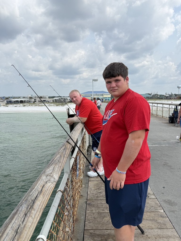Damon standing on a pier with a fishing pole while his dad leans against the railing behind him.