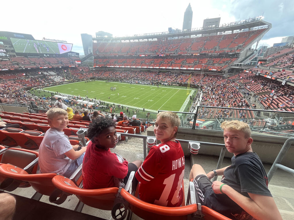 Four students sitting in stadium seats, turned around and smiling with the football stadium in the background