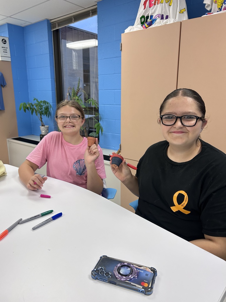 Two students sitting together at a table smiling while holding markers.