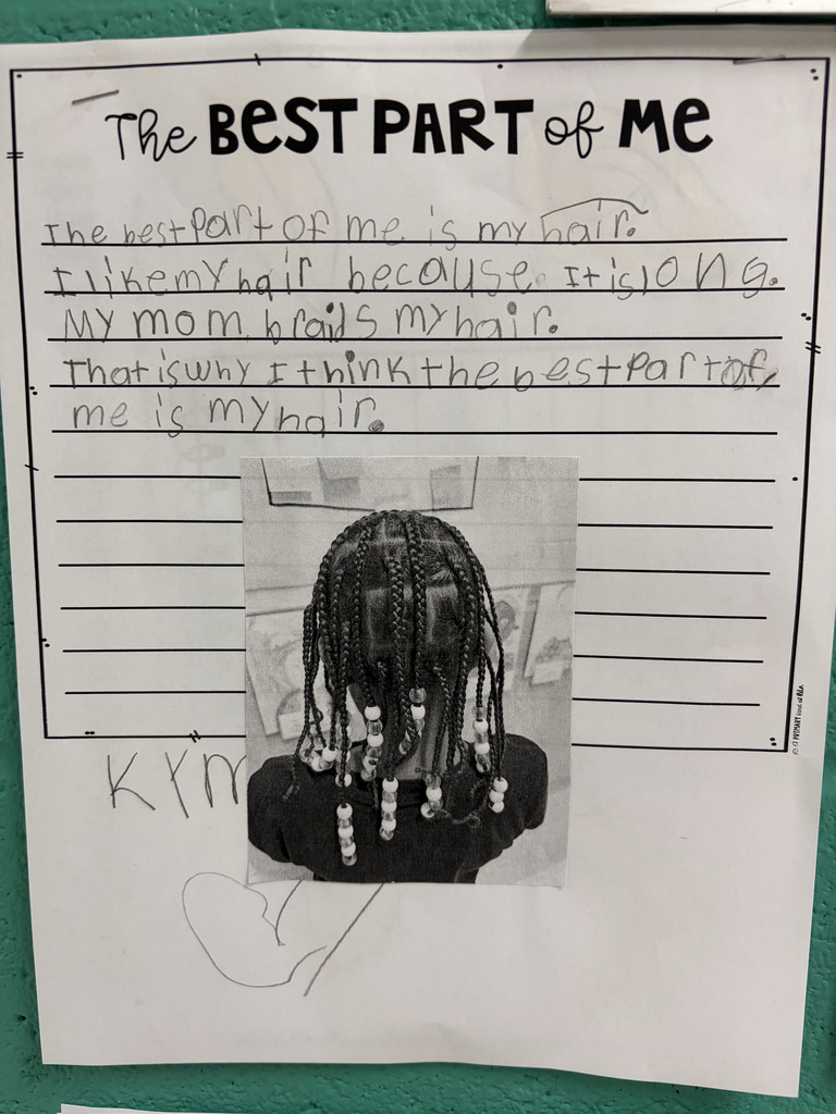 A student worksheet titled “The Best Part of Me” is displayed on a classroom wall. The student wrote about why her hair is the best part of her. A black-and-white photo at the bottom shows the back of the student’s head, highlighting her braided hair decorated with beads.  The student’s writing reads: “The best part of me is my hair. I like my hair because it is long. My mom braids my hair. That is why I think the best part of me is my hair.”