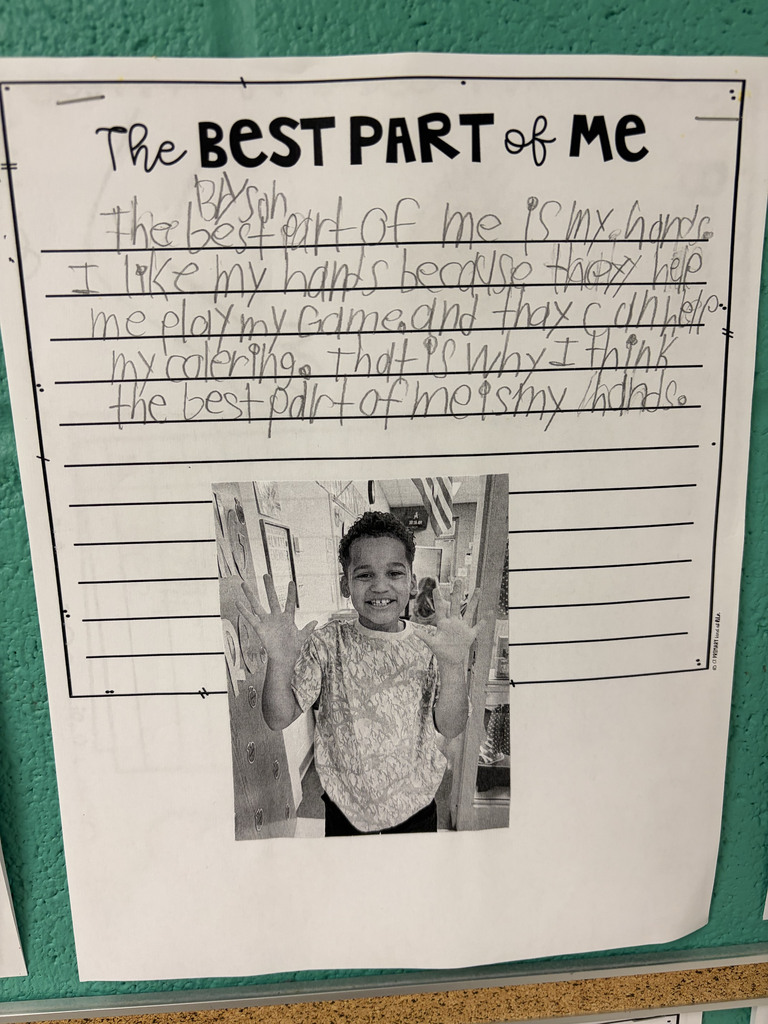 A student worksheet titled “The Best Part of Me” is displayed on a classroom wall. The student, Bryson, wrote about why his hands are the best part of him. A black-and-white photo at the bottom shows the smiling student holding both hands up toward the camera.  The student’s writing reads: “The best part of me is my hands. I like my hands because they help me play my game and they can help my coloring. That is why I think the best part of me is my hands.”