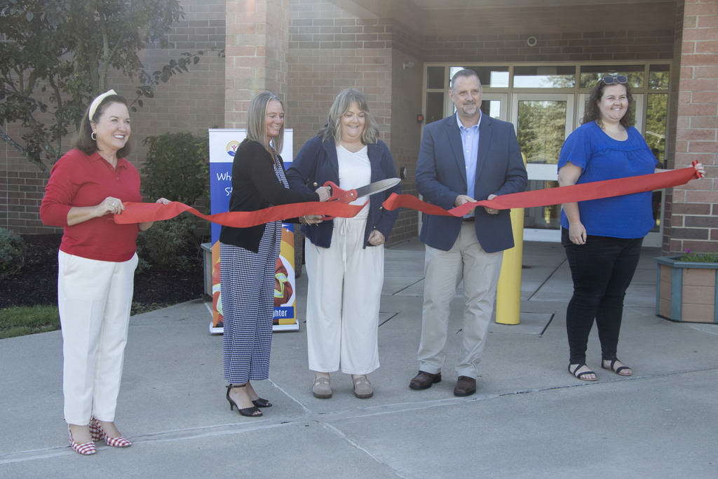 5 people hold a large ceremonial ribbon in front of Alliance Elementary School as the center person uses a pair of big scissors to cut the ribbon