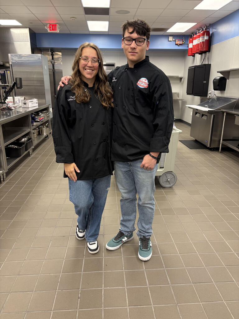 A teacher and a student wearing chef jackets while standing in the culinary arts kitchen