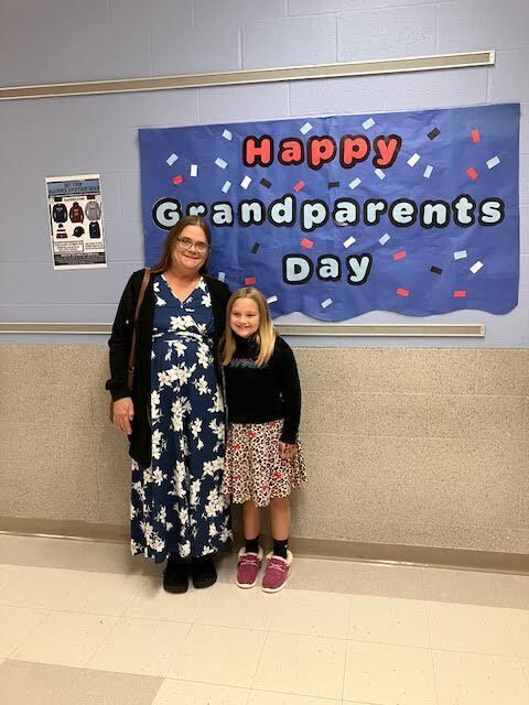 A grandmother and granddaughter stand in front of a fun banner that reads happy grandparents day