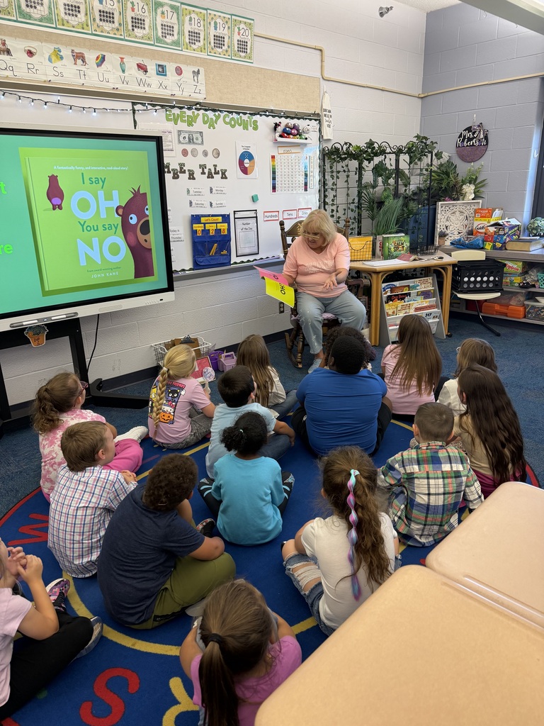 Mrs. Skidmore sitting in the front of the class room with the students sitting around her while she reads a book