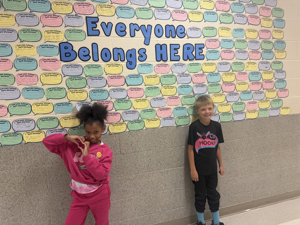 Two students standing infront of a wall covered in cut out conversation bubbles surrounding the words "Everyone belongs here"