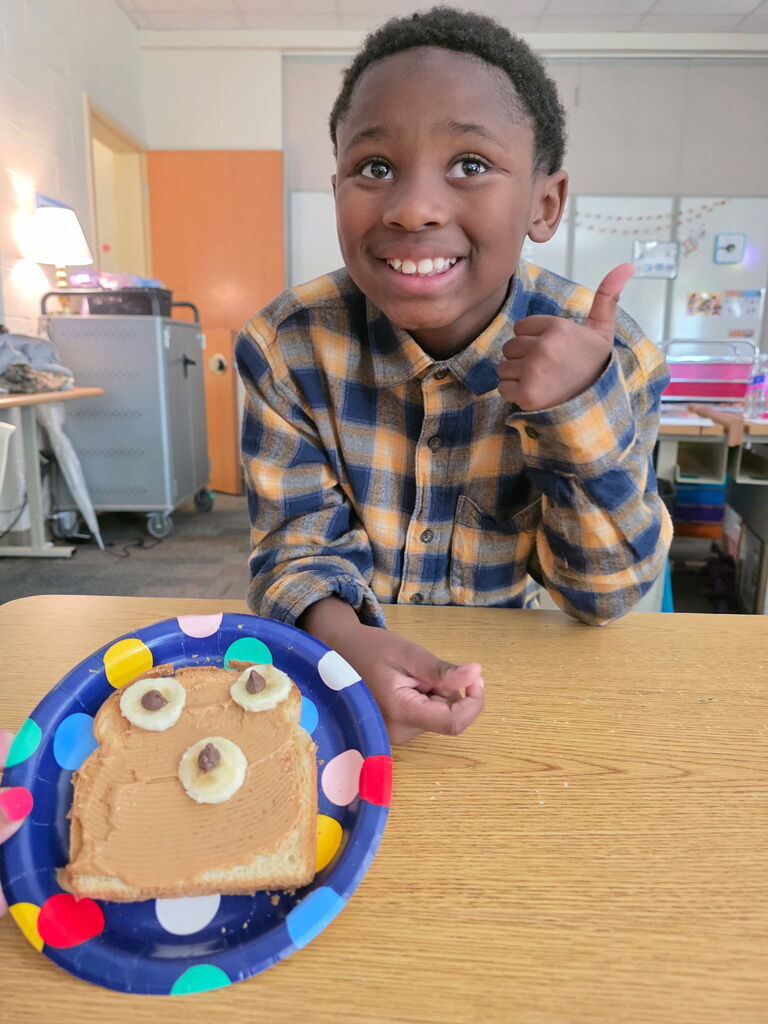 A student gives a thumbs up while a plate sits beside him on the table with a piece of toast that has peanut butter, banana slices and chocolate chips made to look like a bears face