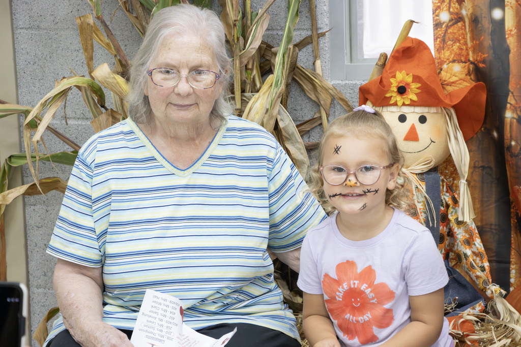 A granddaughter with her face painted like a scarecrow smiles while sitting beside her grandmother on a haybail