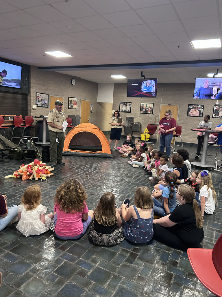 students sit in a circle around a fake campfire with a tent in the background while a troop leader speaks to the students 