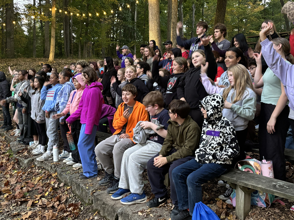 A large group of students sit on wooden bleachers