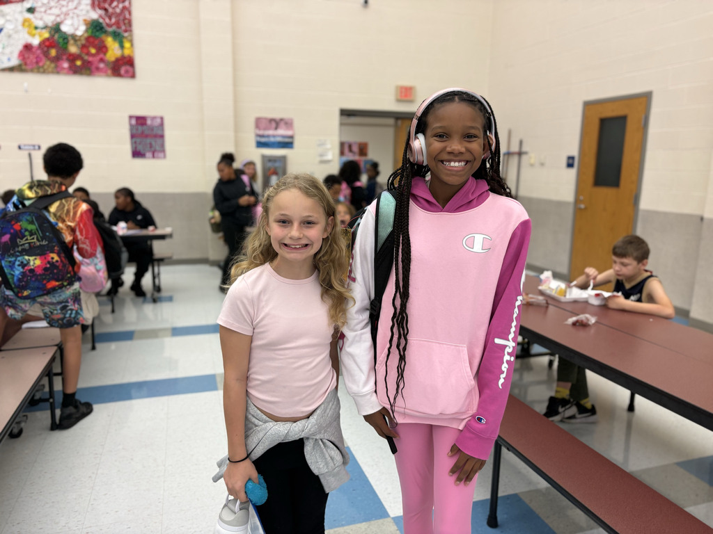 Two students stand together and smile in the lunch room. Both are wearing pink outfits