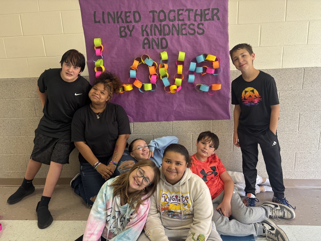 7 students standing in front of a purple banner with the words "Linked together by kindness and love" The word love is made out of neon paper chains